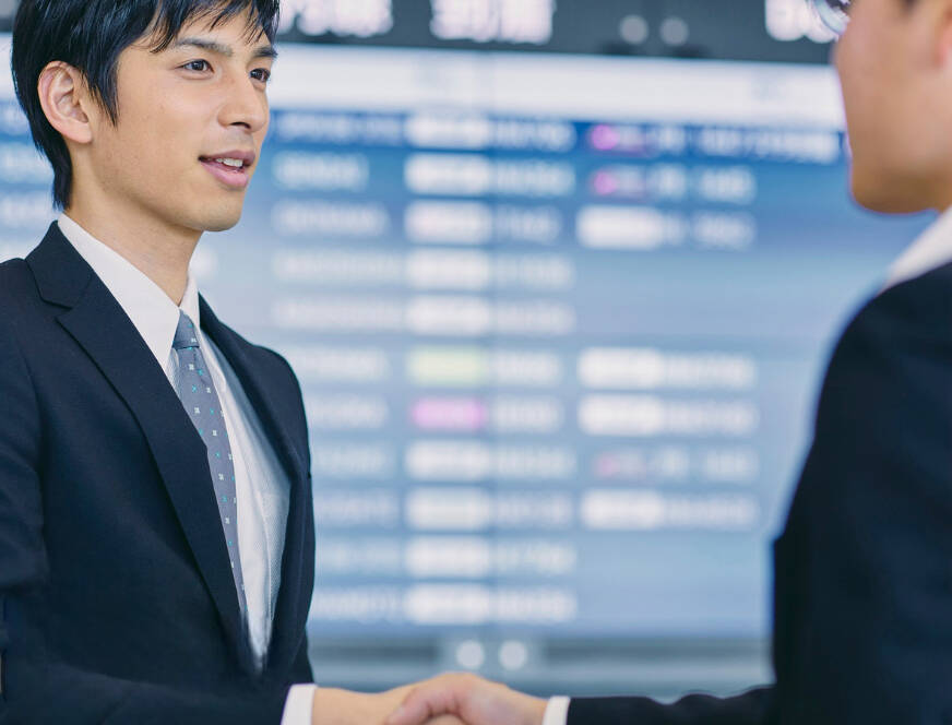 ALT Text: Professional airport greeter in a suit welcoming a client with a handshake in front of a digital arrivals board