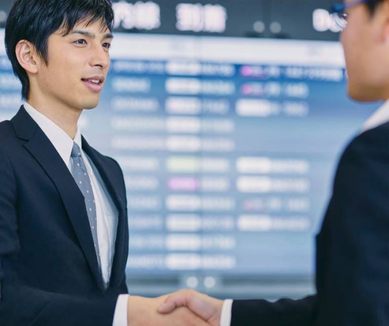 ALT Text: Professional airport greeter in a suit welcoming a client with a handshake in front of a digital arrivals board